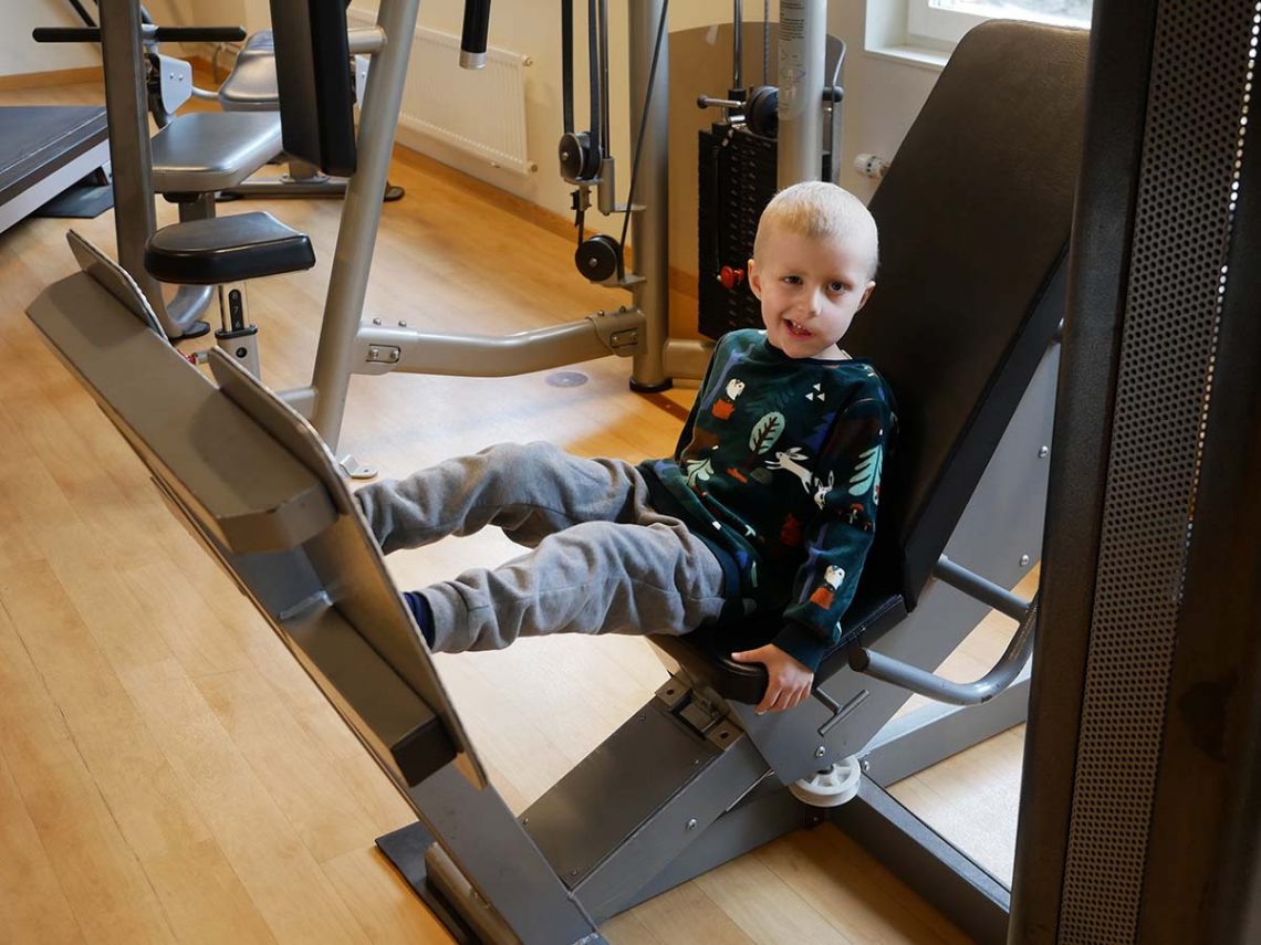 En leende Elis testar benpressen på brandmännens gym