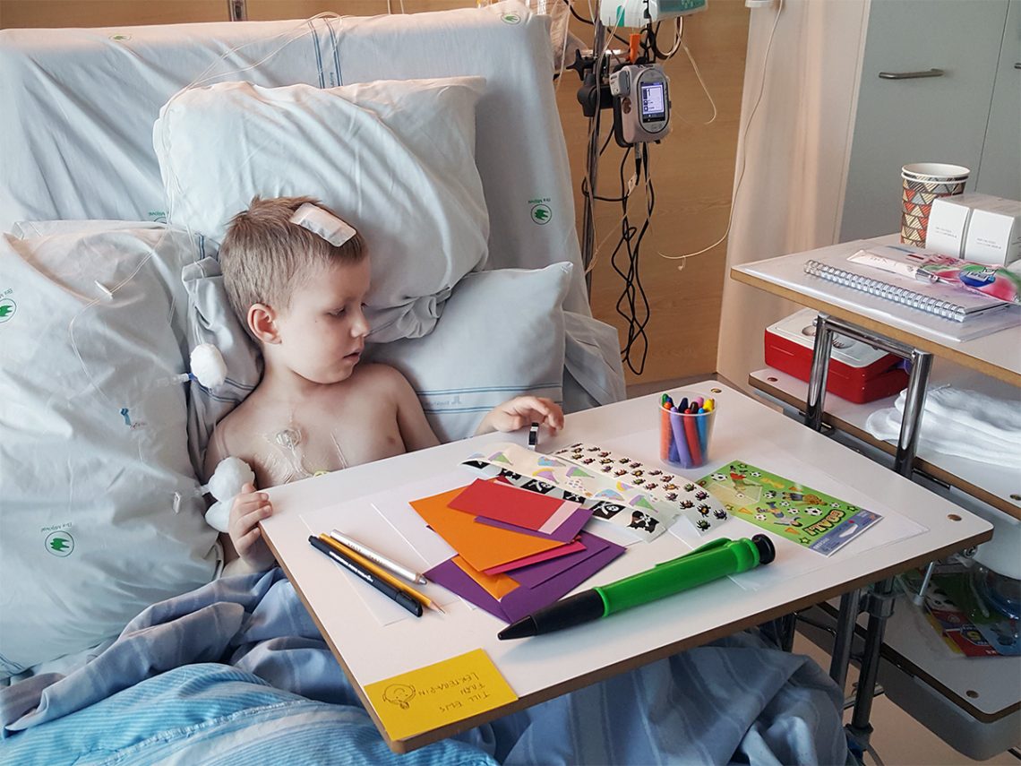 Elis i sjukhussängen, med slangar och kompresser, tittar på pysselsaker på bordet framför honom