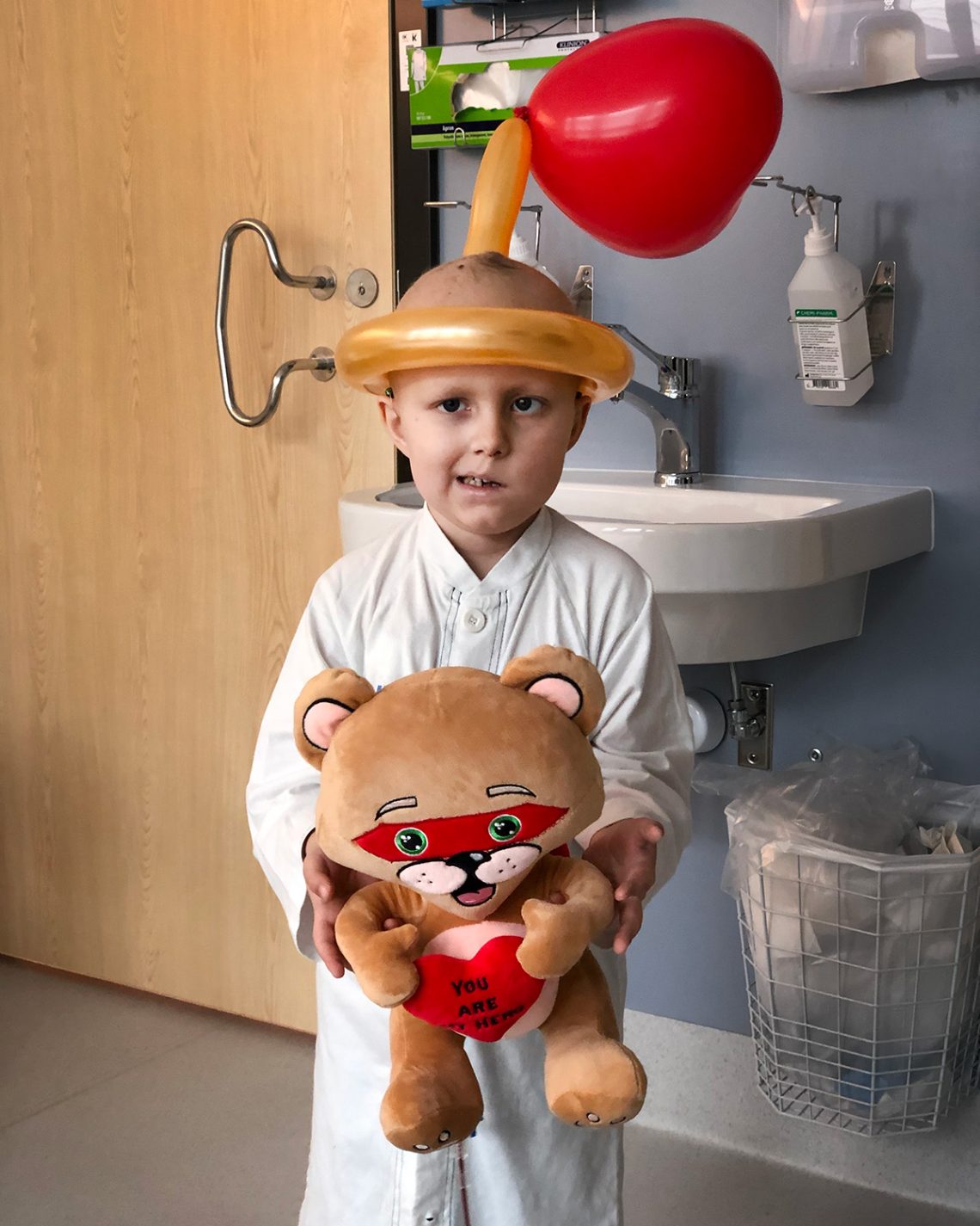 Elis i sjukhusskjorta med en Alla Hjärtans-nalle i famnen, och en hjärt-huvudbonad av ballonger.