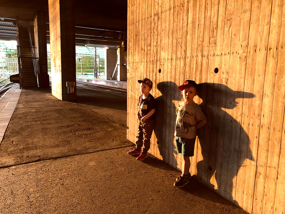Elis och Folke lutar sig mot en beetongvägg på Centralen medan kvällssolen gör deras skuggor långa.