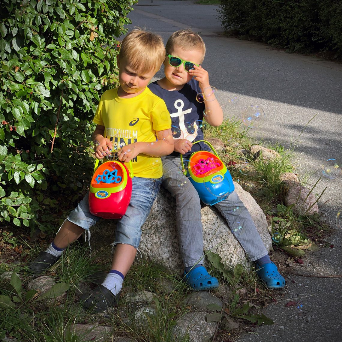 Folke och Elis sitter på en sten med varsin såpbubbelmaskin i händerna