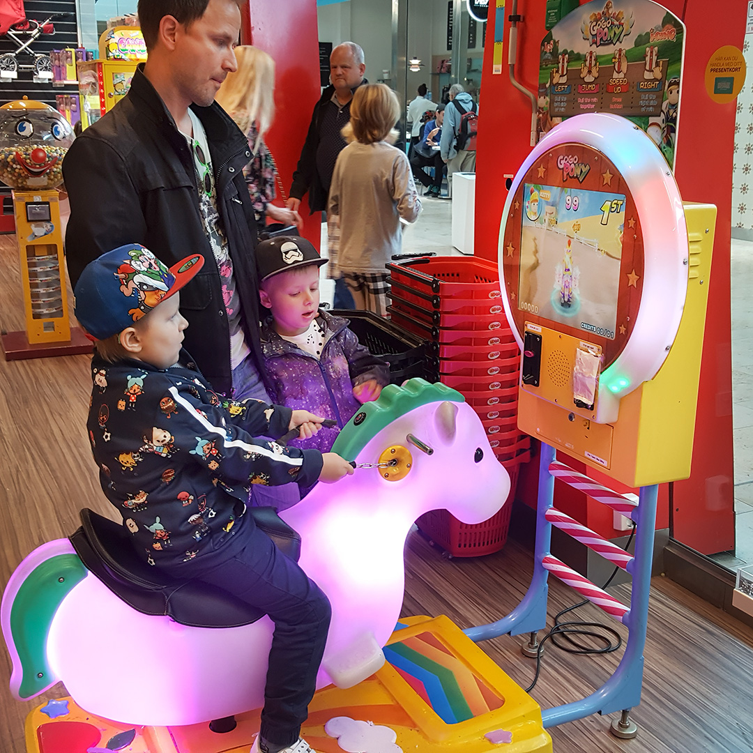 Elis kör ett arkadspel sittande på en gungande häst i Täby centrum, med pappa och Folke som tittar på