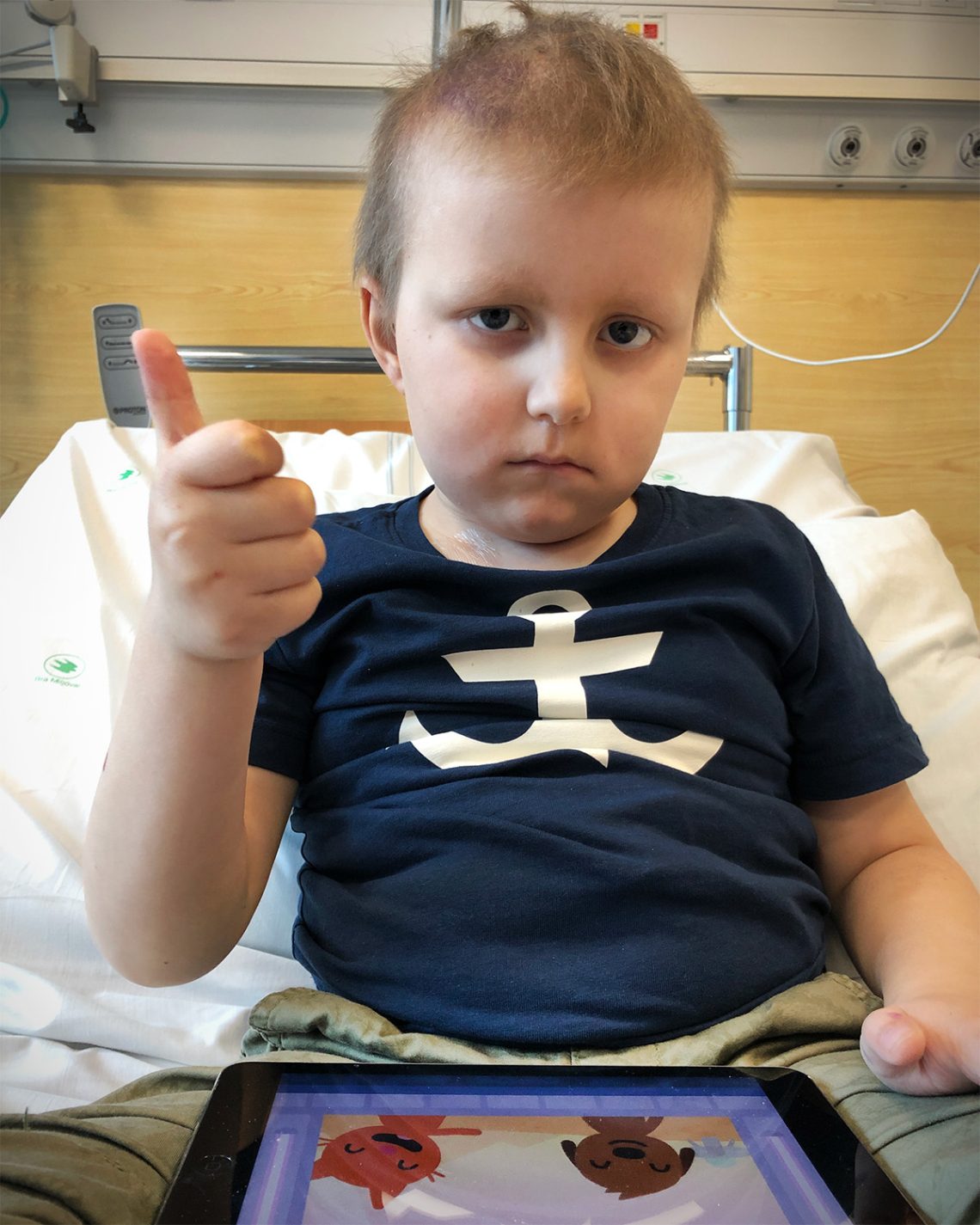 Elis gör tummen upp medan han sitter i sjukhussängen och spelar Sago Mini på iPaden