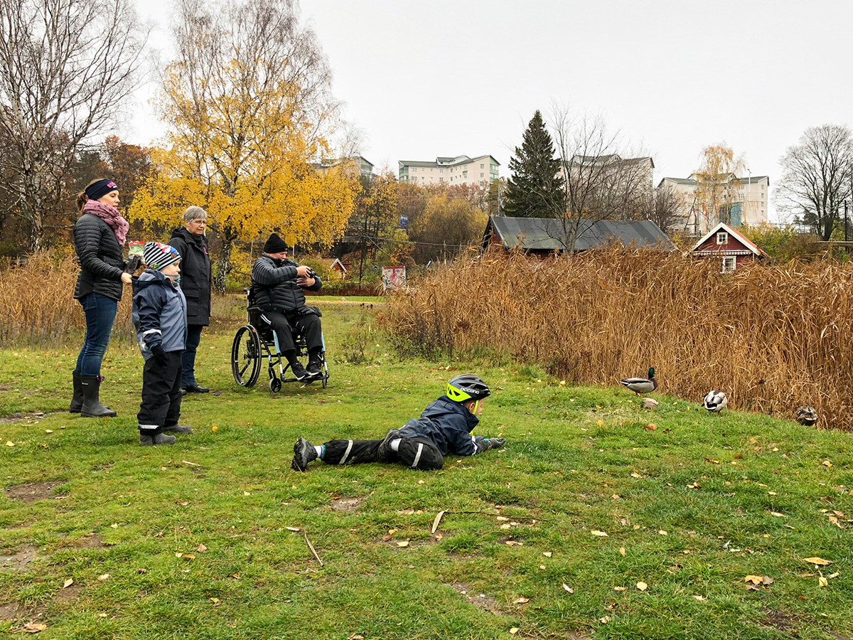 Mamma, Elis, Folke, mormor och morfar spanar på änder. Folke kryper och morfar i rullstolen fotograferar.