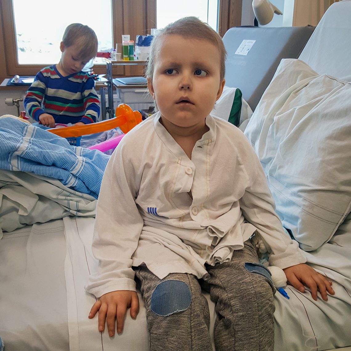 Elis sitter i sjuksängen med Folke i bakgrunden.